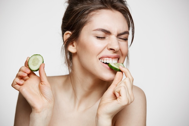 Ragazza nuda naturale abbastanza giovane con pelle pulita perfetta che mangia la fetta del cetriolo sopra fondo bianco. Trattamento facciale.