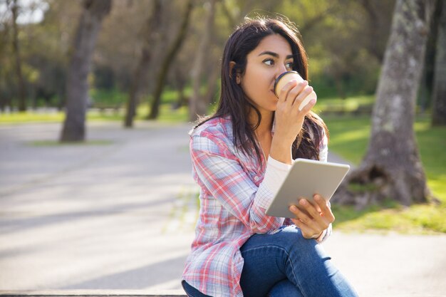 Ragazza pacifica dei pantaloni a vita bassa che gode del ebook e del caffè di mattina in parco