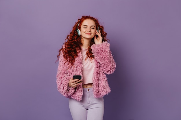 Ragazza pacificata in vestito rosa e bianco alla moda che gode della musica in cuffie blu. Donna dai capelli rossi in posa con lo smartphone sullo spazio lilla.