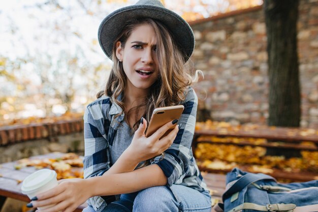 Ragazza pallida preoccupata con i capelli lunghi ricci che guardano la telecamera che tiene il telefono su sfondo autunnale. Modello femminile emotivo in camicia a scacchi beve caffè e legge messaggi.