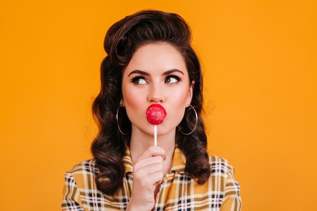 Ragazza pensierosa con l'acconciatura elegante che lecca la caramella. Studio shot di pinup donna con lecca-lecca isolato su sfondo giallo.