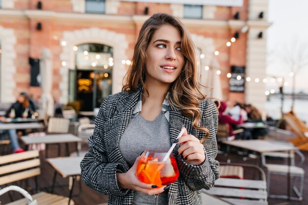 Ragazza pensierosa in elegante cappotto grigio guardandosi intorno, mentre aspetta qualcuno nel caffè all'aperto