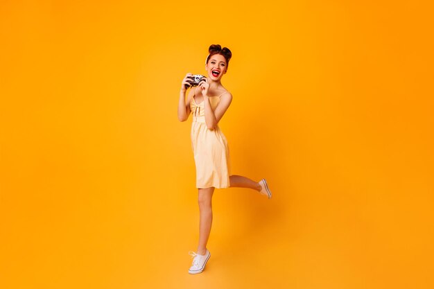 Ragazza pinup emotiva con fotocamera che balla su sfondo giallo Foto in studio di una fotografa in abito