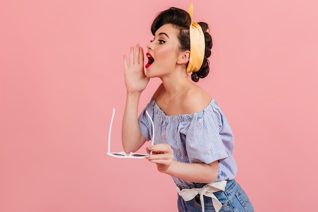 Ragazza pinup in camicetta a righe che grida a qualcuno. Bella donna dai capelli castani con occhiali da sole in posa su sfondo rosa.