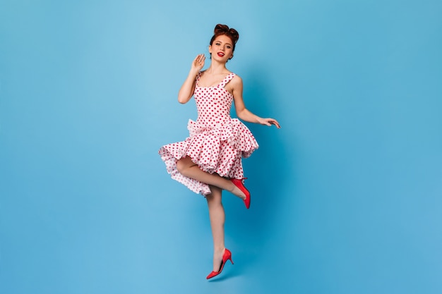Ragazza pinup ispirata con i capelli rossi in piedi su una gamba. Studio shot di donna elegante in abito a pois ballando sullo spazio blu.
