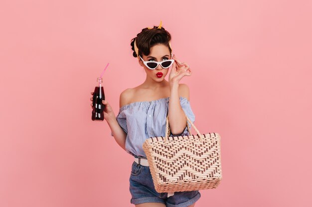 Ragazza pinup pensierosa con una bottiglia di soda toccando occhiali da sole. Studio shot di raffinata donna elegante isolata su sfondo rosa.