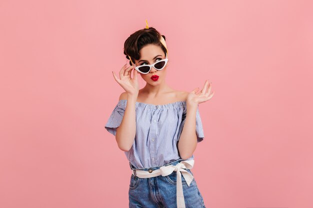 Ragazza pinup pensierosa in occhiali da sole in piedi su sfondo rosa. Studio shot di bruna giovane signora in abito estivo.