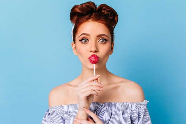 Ragazza pinup sorpresa che lecca caramelle dure Vista frontale di una donna caucasica emotiva con lecca-lecca isolata su sfondo blu