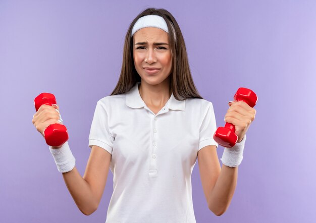 Ragazza piuttosto sportiva dispiaciuta che indossa la fascia e il braccialetto che tengono i manubri isolati sullo spazio viola