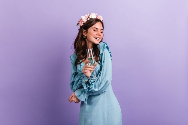 Ragazza positiva di buon umore ride mentre si gode la festa sul muro viola. Modello in vestito blu che tiene un bicchiere di champagne.