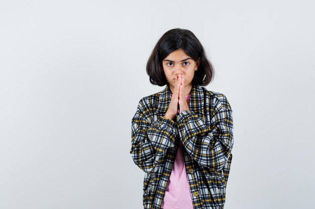 Ragazza preadolescente in camicia, giacca che tiene le mani unite sulla bocca mentre si aspetta notizie e sembra paziente, vista frontale.