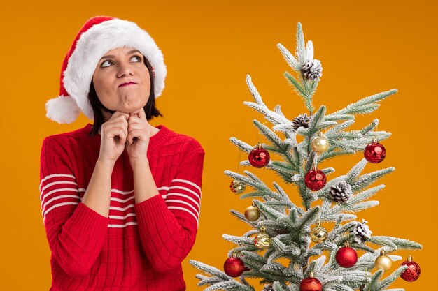 Ragazza premurosa che porta il cappello della santa che sta vicino all'albero di Natale decorato che osserva in su tenendo le mani sotto il mento con le labbra increspate isolate sulla parete arancione