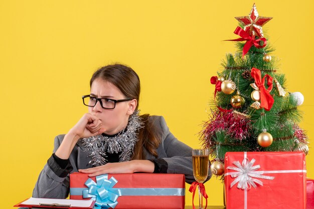 Ragazza premurosa di vista frontale con gli occhiali che si siedono al tavolo pensando vicino all'albero di Natale e al cocktail dei regali