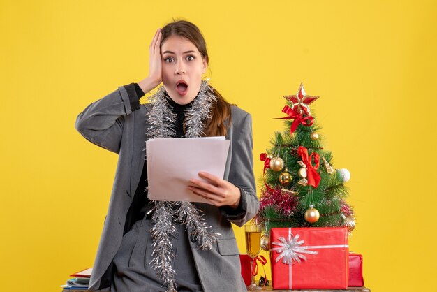 Ragazza premurosa di vista frontale con i documenti che tengono la sua testa vicino all'albero di Natale e al cocktail dei regali