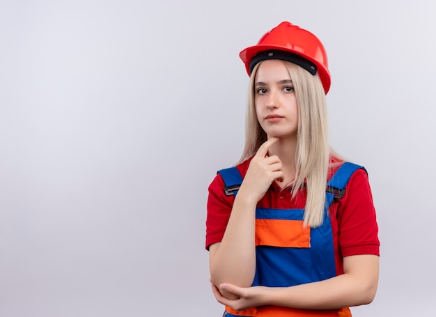 Ragazza premurosa giovane bionda ingegnere costruttore in uniforme che mette il dito sul mento sulla parete bianca isolata con lo spazio della copia