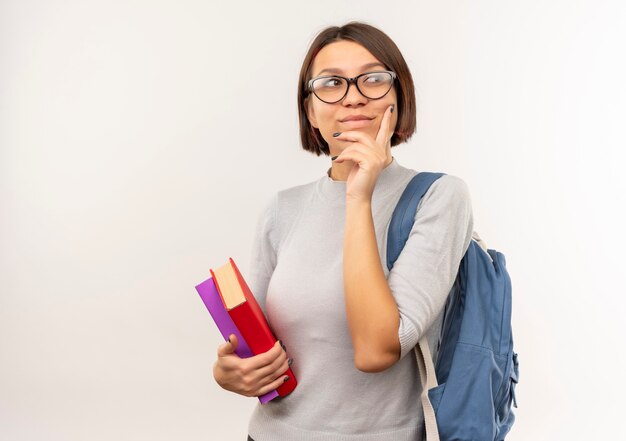 Ragazza premurosa giovane studente con gli occhiali e borsa posteriore che tengono i libri mettendo la mano sul mento isolato sul muro bianco