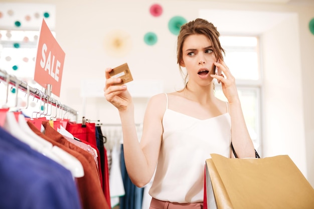 Ragazza premurosa in cima bianca in piedi vicino a appendiabiti in vendita e guardando stupita la sua carta di credito in boutique. Ritratto di signora che parla sul suo cellulare con borse della spesa in mano nel negozio di vestiti