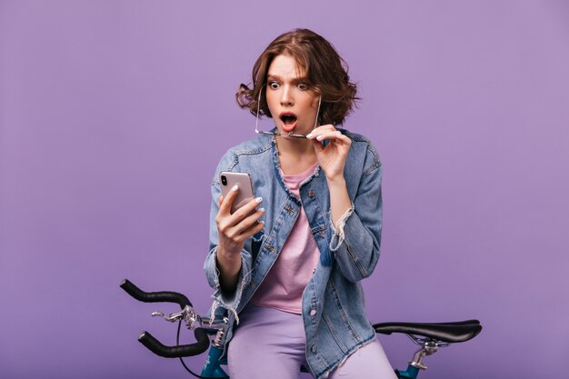 Ragazza preoccupata in giacca di jeans guardando lo schermo del telefono. Tiro al coperto di attraente signora caucasica che si siede sulla bicicletta con lo smartphone e che esprime stupore.