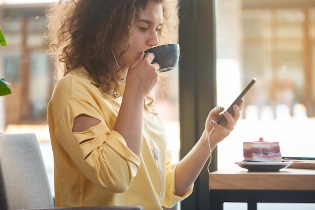 Ragazza riccia che si rilassa bevendo caffè al bar utilizzando smartphone