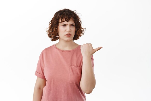 Ragazza riccia delusa e confusa, che punta a destra con la faccia accigliata scontenta e infastidita, in piedi sconvolta su sfondo bianco, indossa una t-shirt estiva