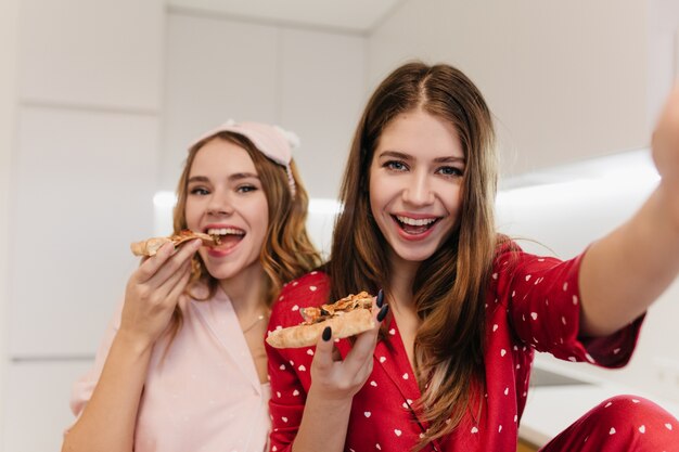 Ragazza riccia emotiva che mangia pizza con il sorriso. Foto interna della donna dai capelli castani felice in indumenti da notte rossi che fa selfie con un amico.