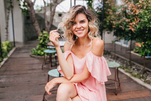 Ragazza romantica che gioca con i capelli corti biondi mentre ci si rilassa nel bellissimo caffè. Attraente donna bionda in posa con l'espressione del viso interessato nel ristorante di strada.