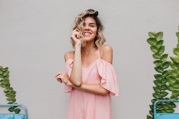 Ragazza romantica con capelli ondulati che tocca il suo fronte con il sorriso. Foto dell'interno della splendida modella abbronzata in piedi vicino alla pianta verde.