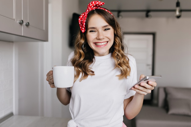 Ragazza romantica di risata con il nastro rosso che gode della mattina con il caffè. Tiro al coperto di adorabile signora riccia che tiene tazza di tè e telefono.