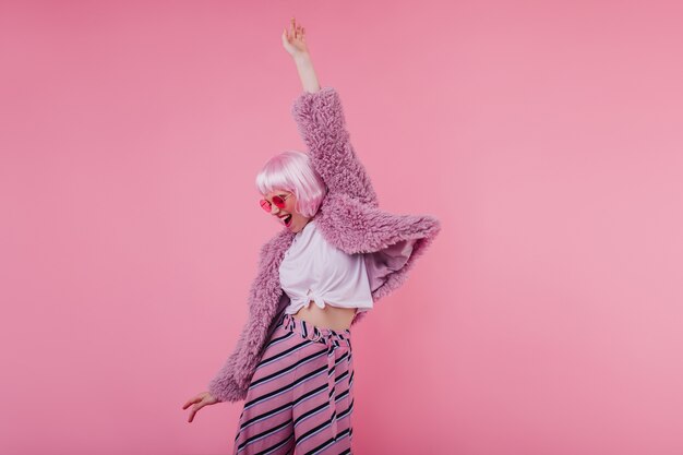 Ragazza romantica in occhiali da sole luminosi e peruke che balla sulla parete rosa. Splendido modello femminile in pantaloni a righe e giacca di pelliccia che ride