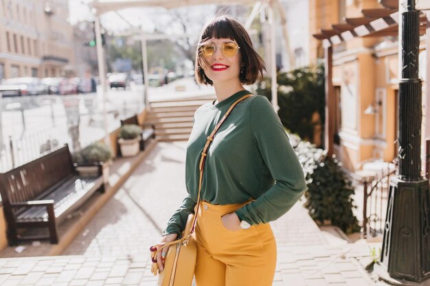 Ragazza romantica ridente in maglione verde che si gode la soleggiata giornata primaverile Splendida donna dai capelli corti con occhiali da sole che si rilassa nel fine settimana