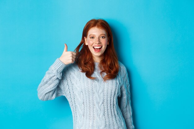 Ragazza rossa allegra in maglione, che mostra il pollice in su in segno di approvazione, come e loda il prodotto, in piedi su sfondo blu.