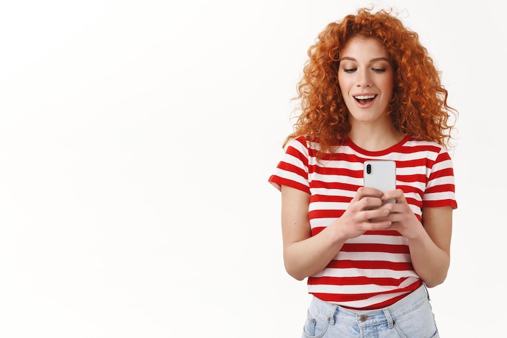 Foto gratuito ragazza rossa che scatta foto amico che registra segretamente video smartphone aspetto giocoso divertendosi sorridente ampiamente sbirciare display telefono scattare foto fresche in piedi sfondo bianco copia spazio