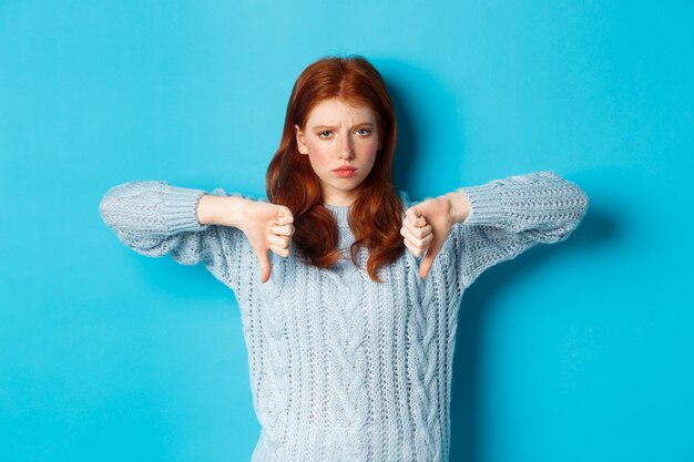 Ragazza rossa delusa in maglione che mostra pollice in giù, giudicando un cattivo prodotto, in disaccordo e antipatia promo, in piedi su sfondo blu