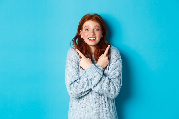 Ragazza rossa eccitata che punta le dita lateralmente, mostrando due scelte e guardando la telecamera tentata, in piedi su sfondo blu.