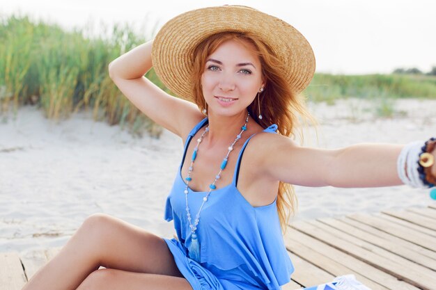 Ragazza rossa felice che fa autoritratto sulla spiaggia. Tenendo il cappello di paglia. Braccialetti e collana alla moda giurando.