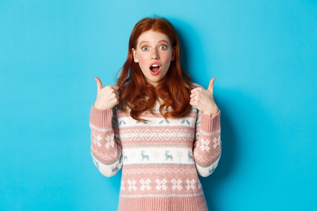 Ragazza rossa impressionata in maglione che mostra pollice in su, bocca aperta affascinata, approva e piace il prodotto, lodando qualcosa, in piedi su sfondo blu.