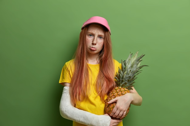 Ragazza rossa lentigginosa e offesa che viene punita dai genitori, tiene un ananas succoso, stringe le labbra e sembra cupa, ha subito un trauma durante lo sport rischioso, posa contro il muro verde.