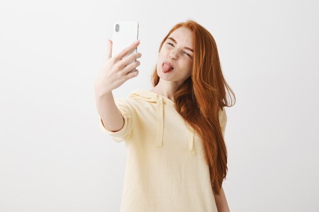 Ragazza rossa sciocca che mostra la lingua e prendendo selfie