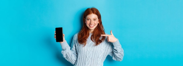 Ragazza rossa soddisfatta che indica lo schermo del telefono che mostra l'app per smartphone o la promozione online e sorride