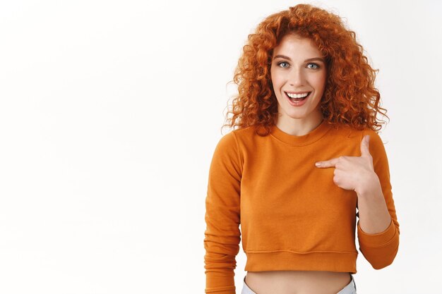 Ragazza rossa sorpresa che indica se stessa chiedendosi se è vincitrice, sorridendo allegramente sorpreso notizie piacevoli, ricevendo un invito festa fresca, sembra impressionato e meravigliato, stando in piedi sul muro bianco