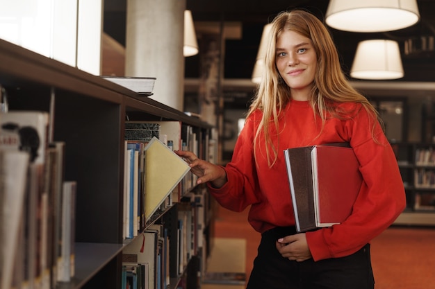 Ragazza rossa, studente di college prende un libro dallo scaffale in biblioteca o libreria, sorridendo alla telecamera.