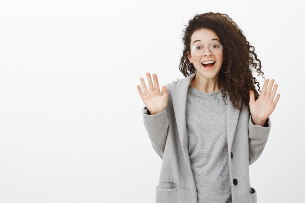 Ragazza sbalordita dalle fantastiche notizie dal bestie. Ritratto di donna europea felice soddisfatta con capelli ricci in occhiali e cappotto grigio, sollevando le palme e sorridendo dalla felicità