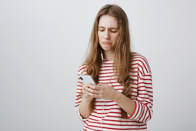 Ragazza sconvolta guardando deluso dallo schermo del telefono cellulare, ascoltando musica in auricolari