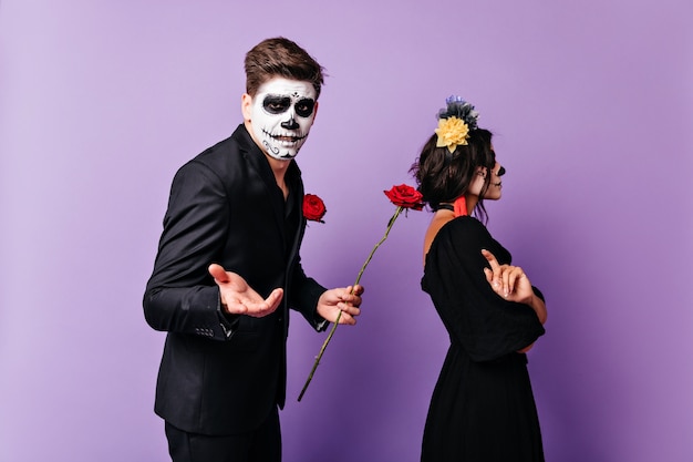 Ragazza sconvolta in costume di carnevale in piedi in studio con il fidanzato. Uomo in abito mascherato che chiede perdono con rose.