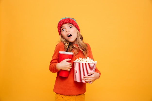 Ragazza scossa in maglione e cappello che tengono popcorn e tazza di plastica mentre esaminando la macchina fotografica sopra l'arancia