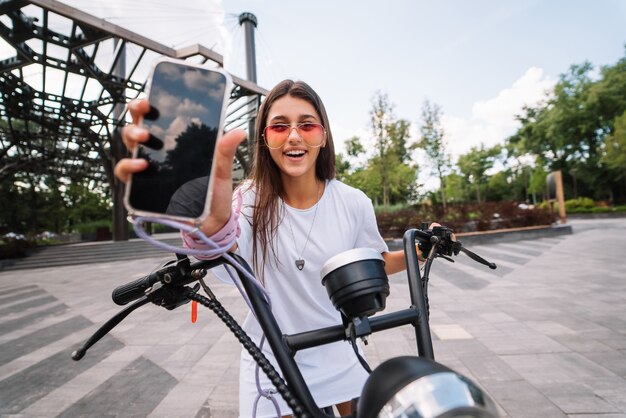 Ragazza seduta su uno scooter e mostra il telefono alla telecamera