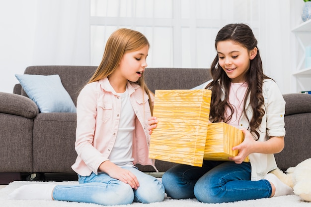 Ragazza seduta sul tappeto guardando la sua amica aprendo la scatola regalo