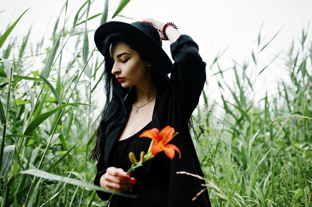 Ragazza sensuale tutta in labbra rosse nere e cappello La donna drammatica gotica tiene il fiore di giglio arancione sulla canna comune