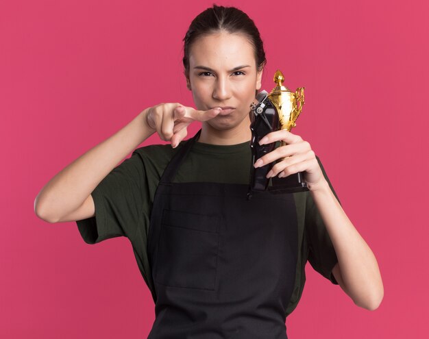 Ragazza seria giovane barbiere bruna in uniforme che tiene tagliacapelli e coppa del vincitore e punta alla telecamera