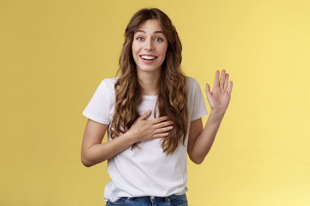 Ragazza si presenta amichevole gioiosa conversazione casuale sorridente ampiamente tenere la mano cuore alzare il palmo agitando giura di dire la verità sii onesto sogghignando fai promessa giuramento stare in piedi sfondo giallo dire ciao